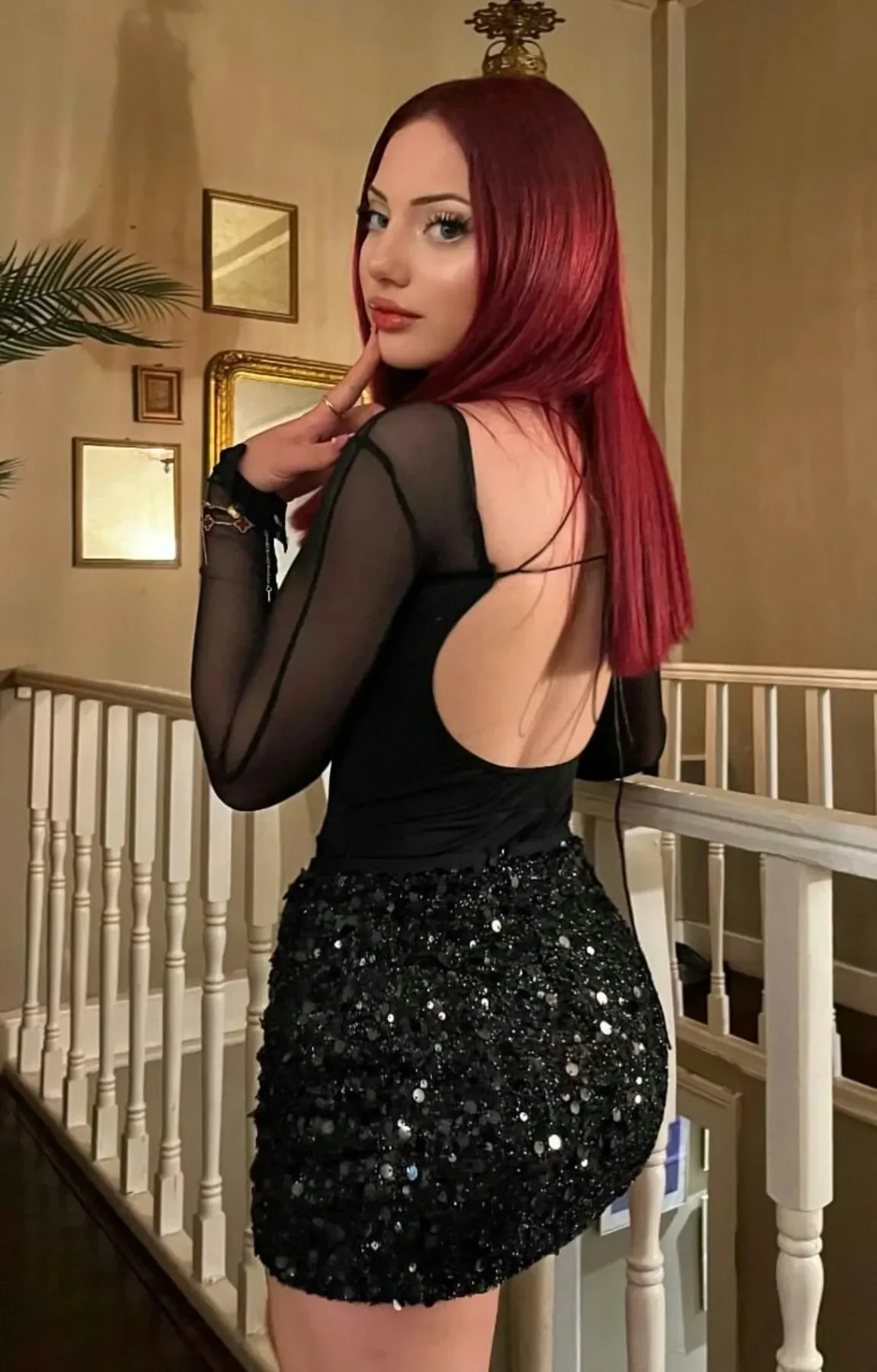 Woman with long red hair posing in a black sequined dress with sheer sleeves indoors, looking over her shoulder.