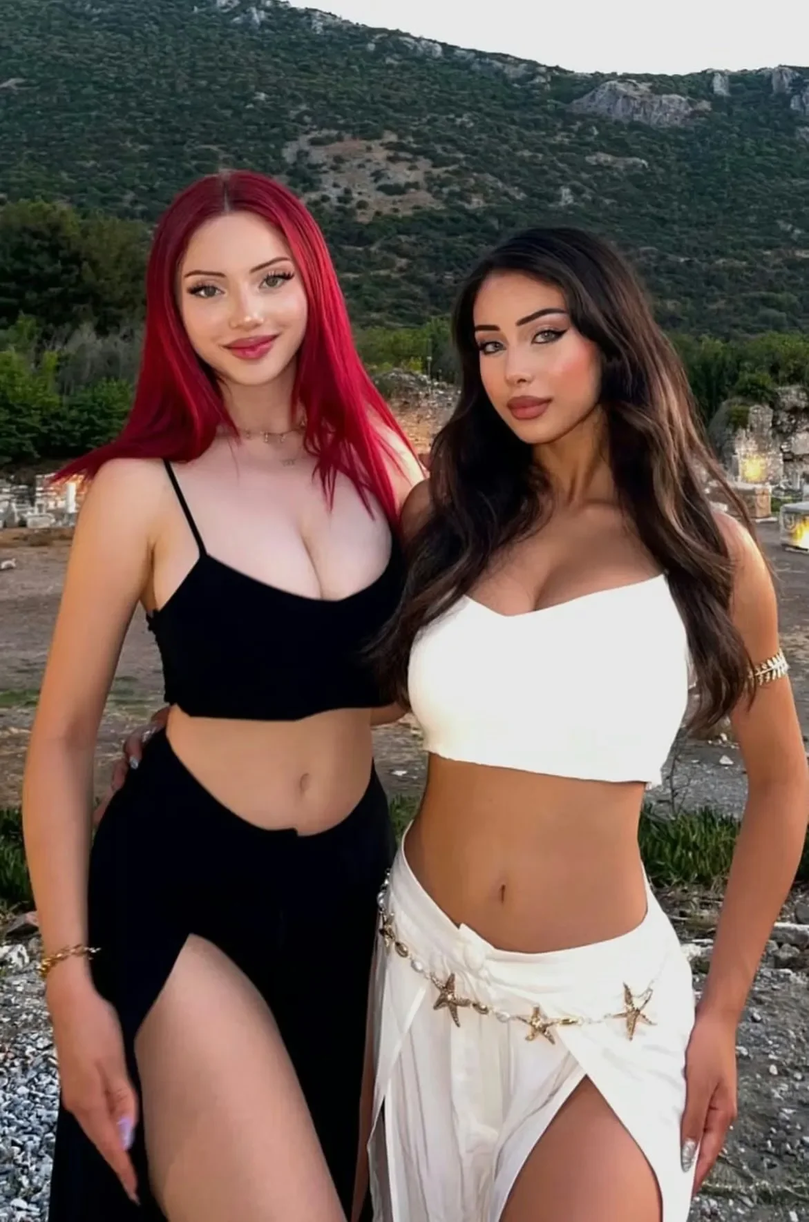 Two young women posing together outdoors in front of a rocky hillside; one with bright red hair wearing a black crop top and skirt, the other with long dark hair wearing a white crop top and a white skirt with gold star accents.