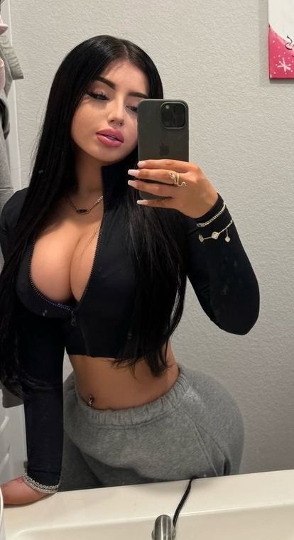 Woman taking a mirror selfie in a bathroom, wearing a black zip-up top and gray sweatpants with long dark hair.