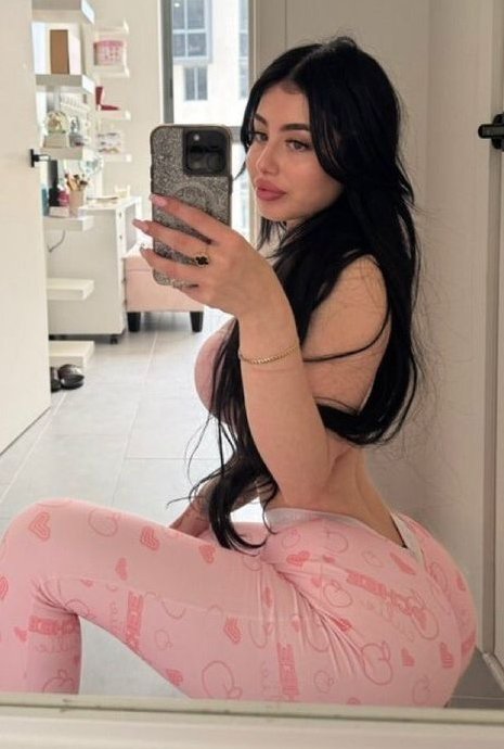 Woman with long black hair taking a mirror selfie in a bright room, wearing pink heart-pattern leggings and a pink top.