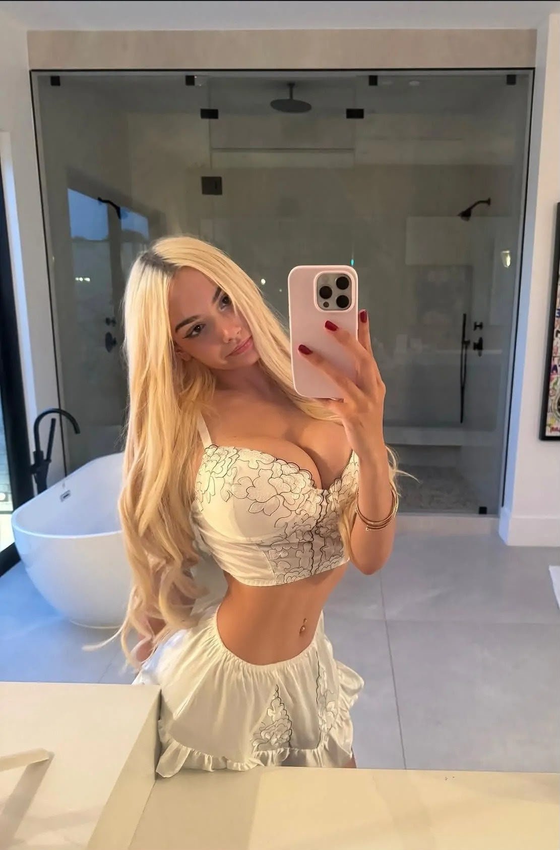 Woman with long blonde hair taking a mirror selfie in a white bathroom, wearing a cream lace crop top and matching skirt.