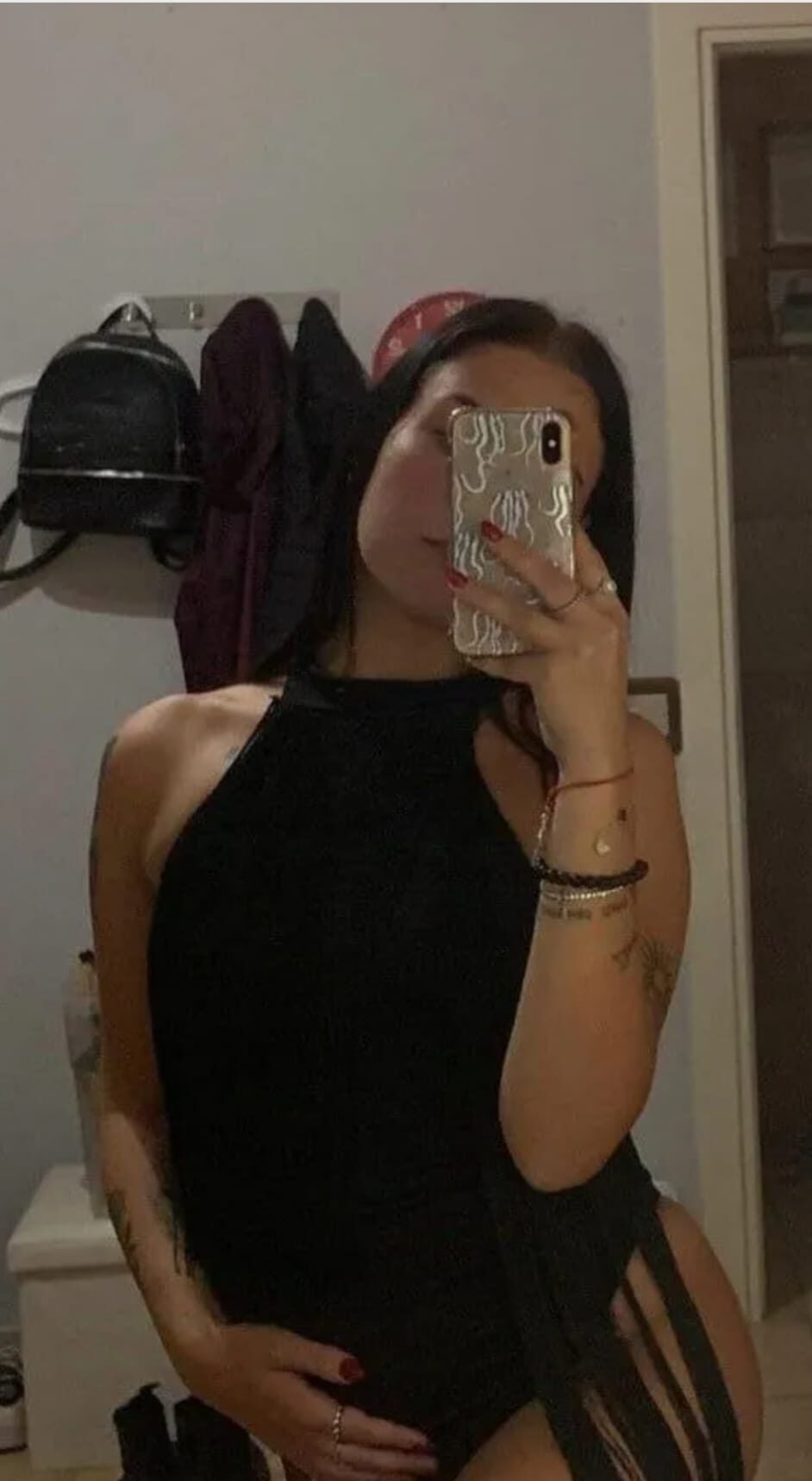 Person in a black halter top taking a mirror selfie, holding a phone with a patterned case; coats hang on a rack behind.