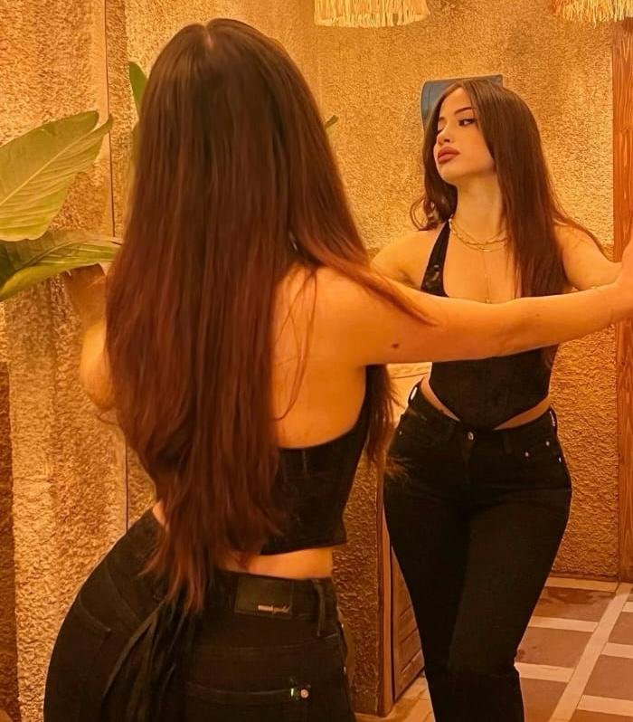 Woman in a black halter top and jeans stands with arms outstretched, looking at her reflection in a mirror under warm lighting.