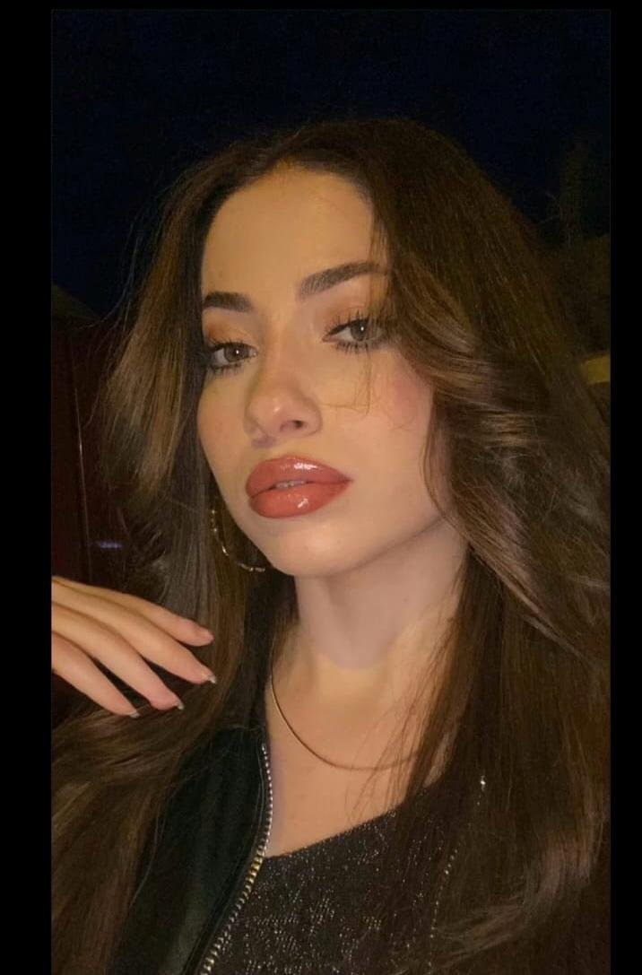 Close-up portrait of a young woman with long brown hair, hoop earrings, and glossy coral lipstick against a dark background.